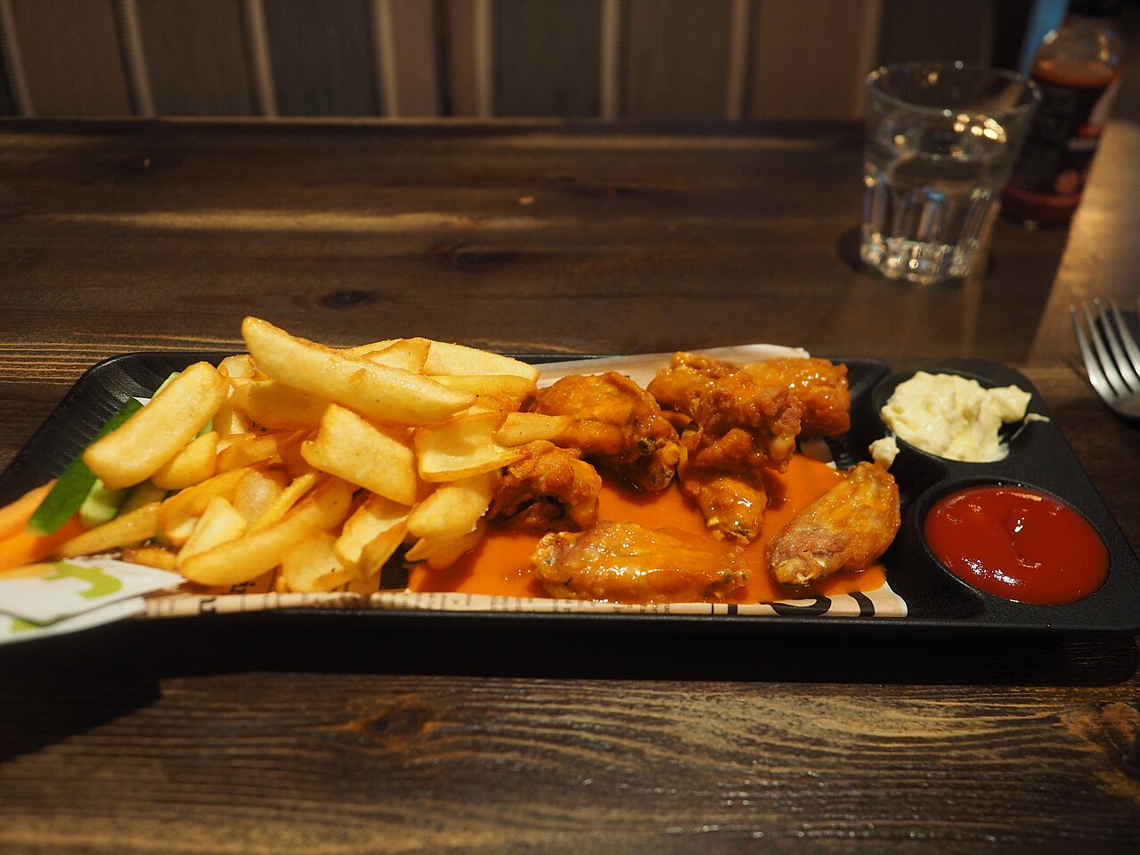 A plate of chicken wings in hot sauce, French fries, carrot sticks and cucumber sticks, along with a glass of water, served for lunch at restaurant West Side Story in Martinlaakso, Vantaa, Finland in 