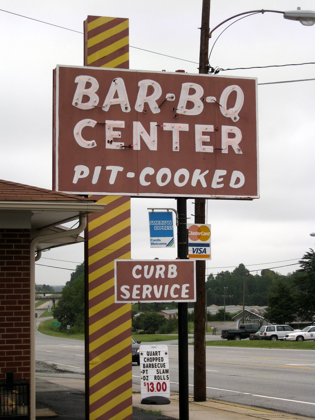 A tall roadside sign on a pylon striped diagonally in maroon and yellow. The brown cabinet carries BAR·B·Q above CENTER in white letters outlined in bare neon tubing, and PIT-COOKED painted in plain white beneath. Below the cabinet hang small panels for American Express, MasterCard and Visa, and a second board reading CURB SERVICE. A plastic A-board on the pavement reads: QUART CHOPPED BARBECUE, PT SLAW, DZ ROLLS, $13.00. The brick corner of the restaurant is at the left and a four-lane road runs behind.