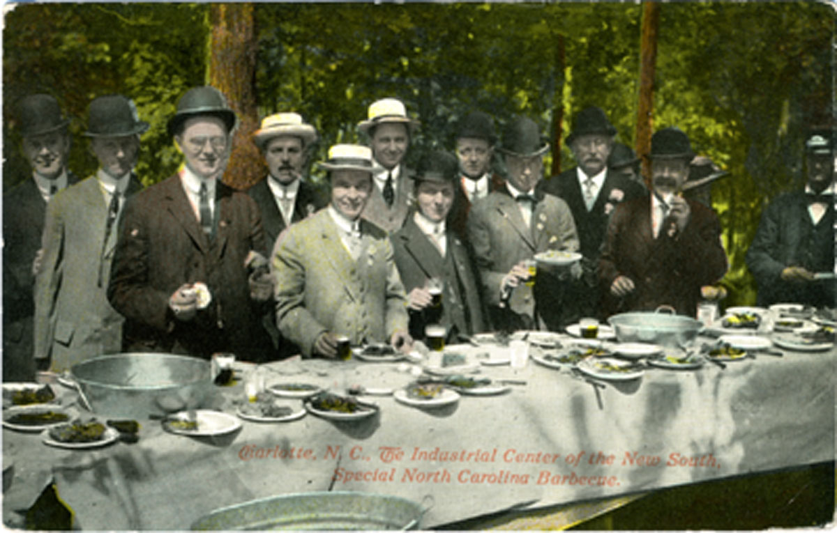 A hand-coloured postcard. Sixteen or so men in suits stand shoulder to shoulder behind a long white-clothed table set up among trees, most in bowler or straw hats, several holding glasses, one a bread roll. The table carries plates of food, tumblers and three big round metal basins. Overprinted across the lower part in red script: “Charlotte, N. C., The Industrial Center of the New South, Special North Carolina Barbecue.”