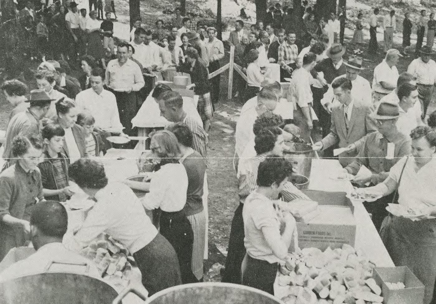 A crowded outdoor serving line, printed as a newspaper halftone so the dot screen shows. Along the near side of long trestle tables, women in aprons and short sleeves serve: one ladles from a metal bucket, another lifts bread rolls out of a big open cardboard carton, a third fills a plate held out to her. Men in shirtsleeves, ties and summer hats file past on the far side with plates and bowls. Behind them the queue winds back uphill among trees past a wooden rail fence, dozens more people standing and sitting on the slope. A galvanised tub stands in the near corner.