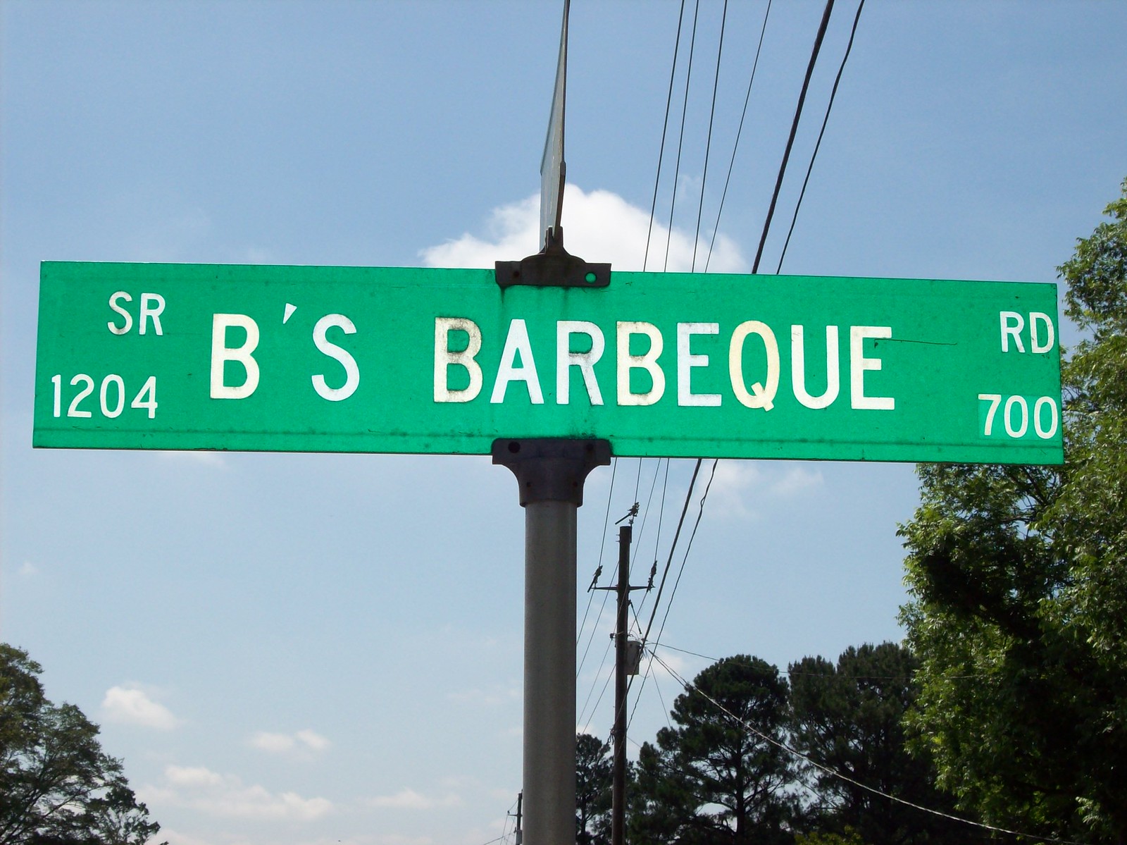 A green street-name blade bolted to a metal post against a blue sky with a few clouds. It reads B'S BARBEQUE in white capitals, with SR 1204 stacked at the left end and RD 700 at the right. Power lines, a utility pole and pine tops fill the lower background.