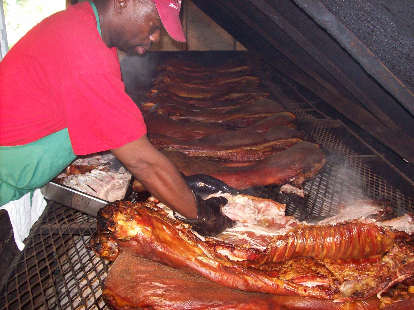 A man in a red T-shirt, green apron and red cap leans into a long open pit and pulls at a cooked hog with a black-gloved hand, opening the ribs and the crisped skin. A row of more cooked hogs recedes down the wire grate behind him, steam rising off them. A steel pan of bones sits on the pit edge by his elbow, a white towel hanging beside it.