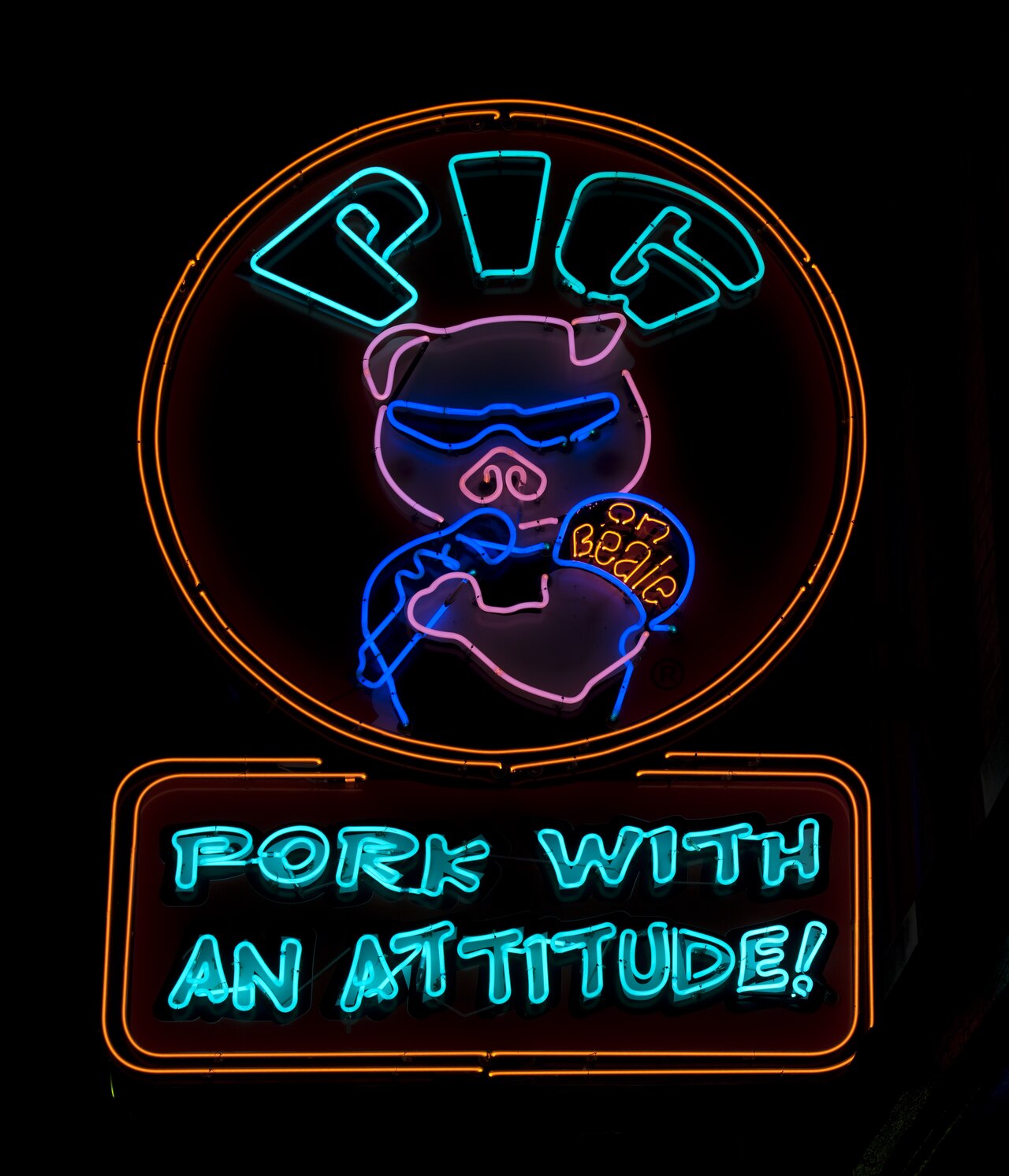 Neon sign at night: a pink pig in blue wraparound sunglasses holds a pork sandwich in both trotters and is biting into it, the word PIG in turquoise above and PORK WITH AN ATTITUDE! on a panel below. The Pig on Beale, Beale Street, Memphis.