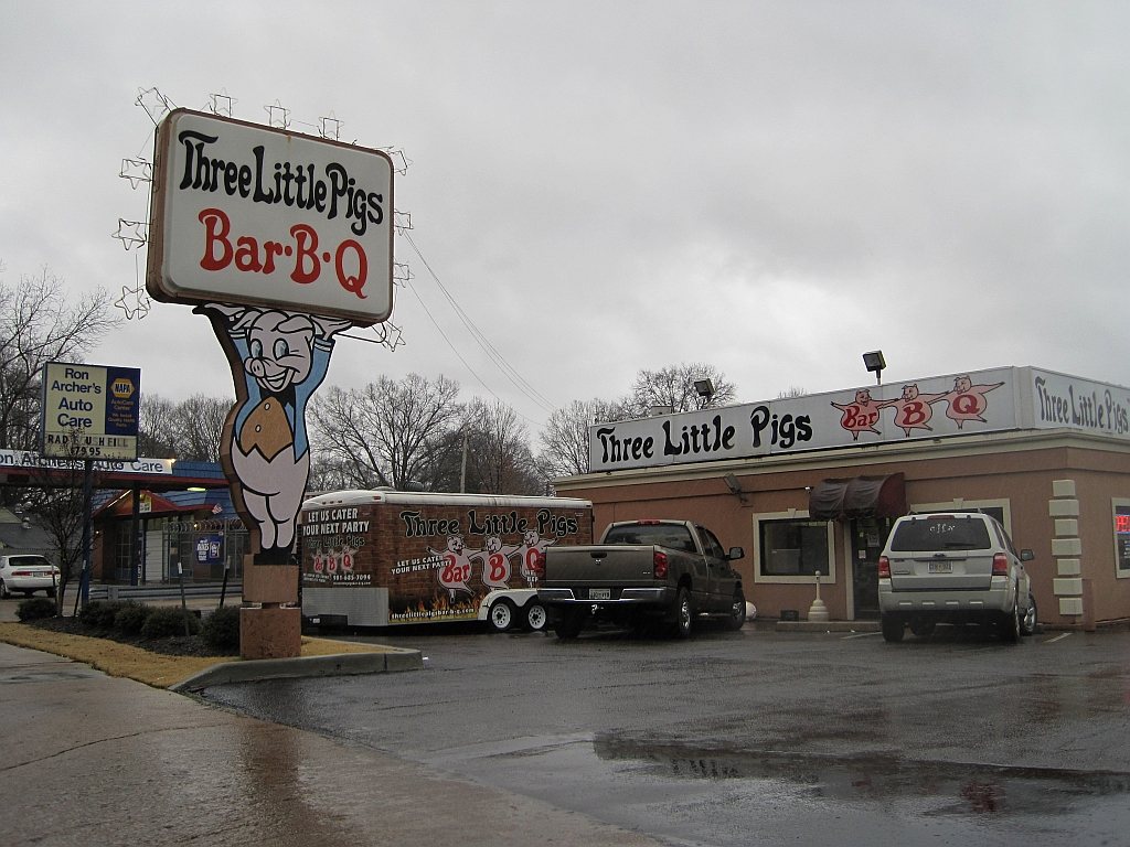 Three Little Pigs Bar-B-Q, Memphis, on a wet January day: a smiling pig in blue dungarees holds the whole signboard over its head at the kerb, and three more pigs along the building's roofline each carry one letter of BAR-B-Q.