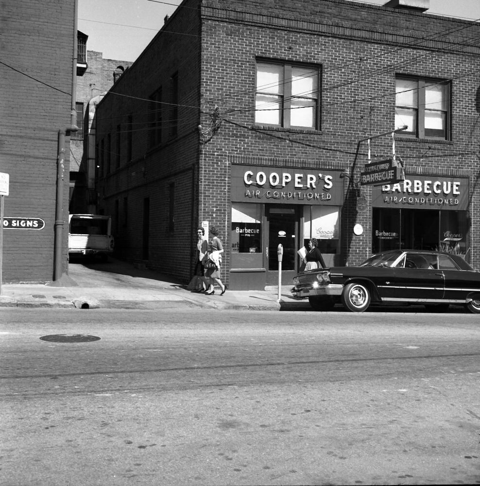 A black-and-white photograph taken from across a downtown street. A two-storey brick building holds the corner; over its ground-floor windows a painted fascia reads COOPER'S, AIR CONDITIONED, and a second awning beside it reads BARBECUE, AIR CONDITIONED, with a hanging bracket sign between them repeating Cooper's Barbecue. Two women walk past the door carrying bags, a long sedan is parked at the kerb and a bus stands in the alley beyond. NO SIGNS is painted on a wall at the left edge.