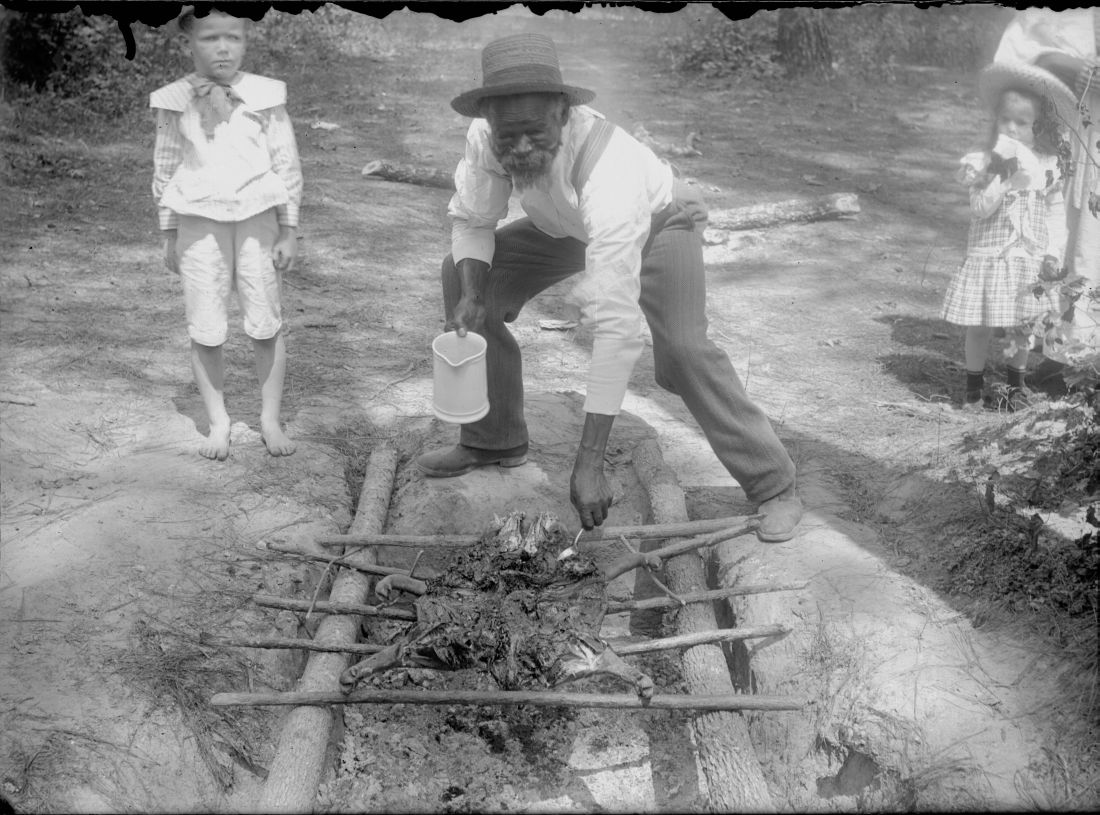 A man in a straw hat, white shirt and braces bends over a shallow trench dug in bare ground. Green poles are laid across the trench as a grate and meat lies on them, smoke rising through. He holds a small tin pail low in his right hand and reaches into the meat with his left. A barefoot boy in a light suit stands at one side of the clearing and a girl in a checked dress and wide hat at the other, both watching. Felled logs and woods behind.