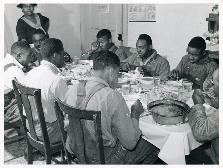 A long table under a white cloth in a farmhouse room, ten or twelve men eating, glasses and plates down its length and a large enamel basin at the near end; a woman stands at the back. The agency caption reads: the Negro tenants and neighbors eating dinner after the white men have finished, at the Wilkins place, Tallyho, Stem, Granville County, November 1939.