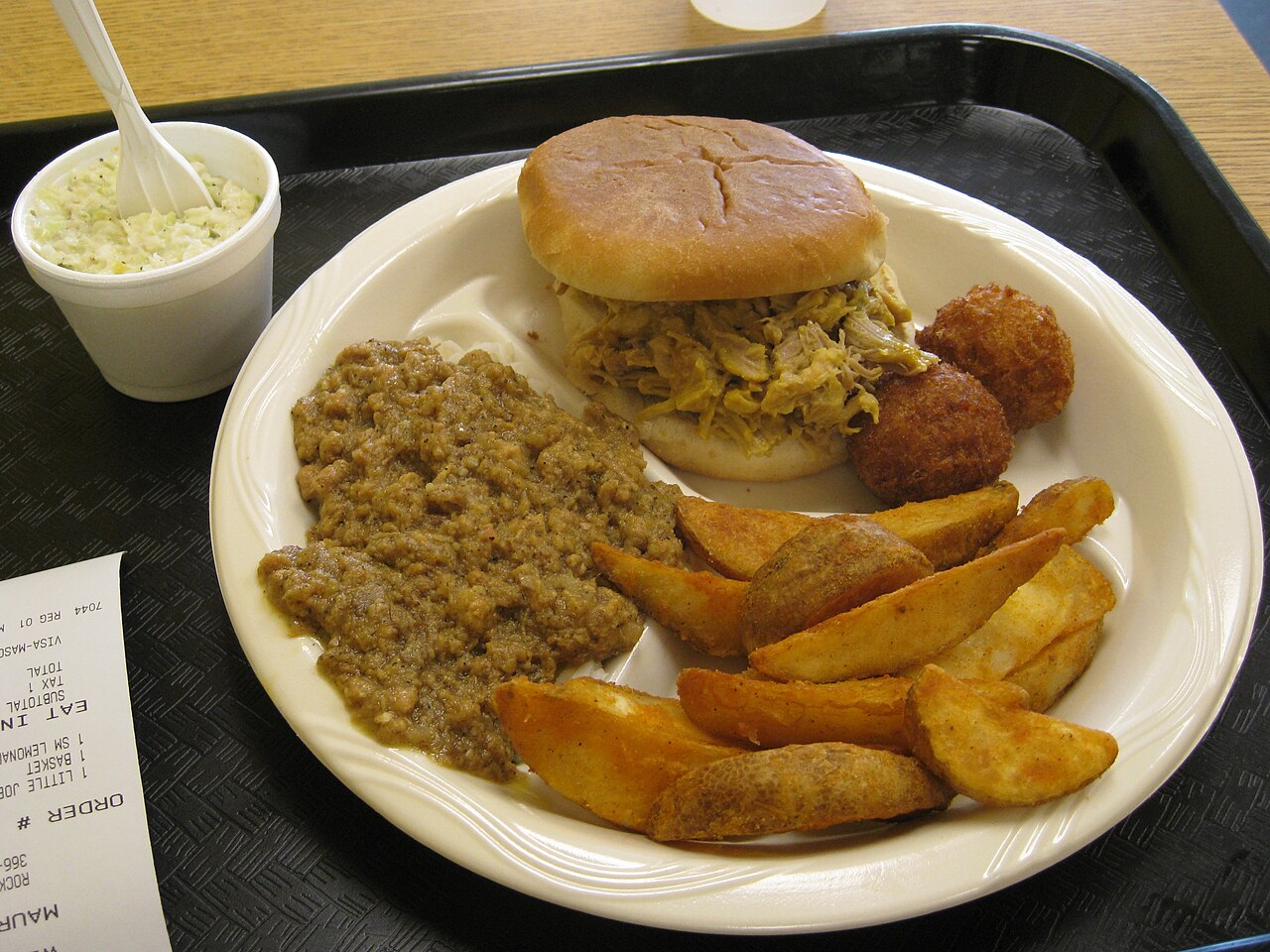 A plate of South Carolina-style barbecue at Maurice's Gourmet BBQ. It includes a pulled pork sandwich, hushpuppies, potato wedges, and hash. A side of coleslaw is in a cup.