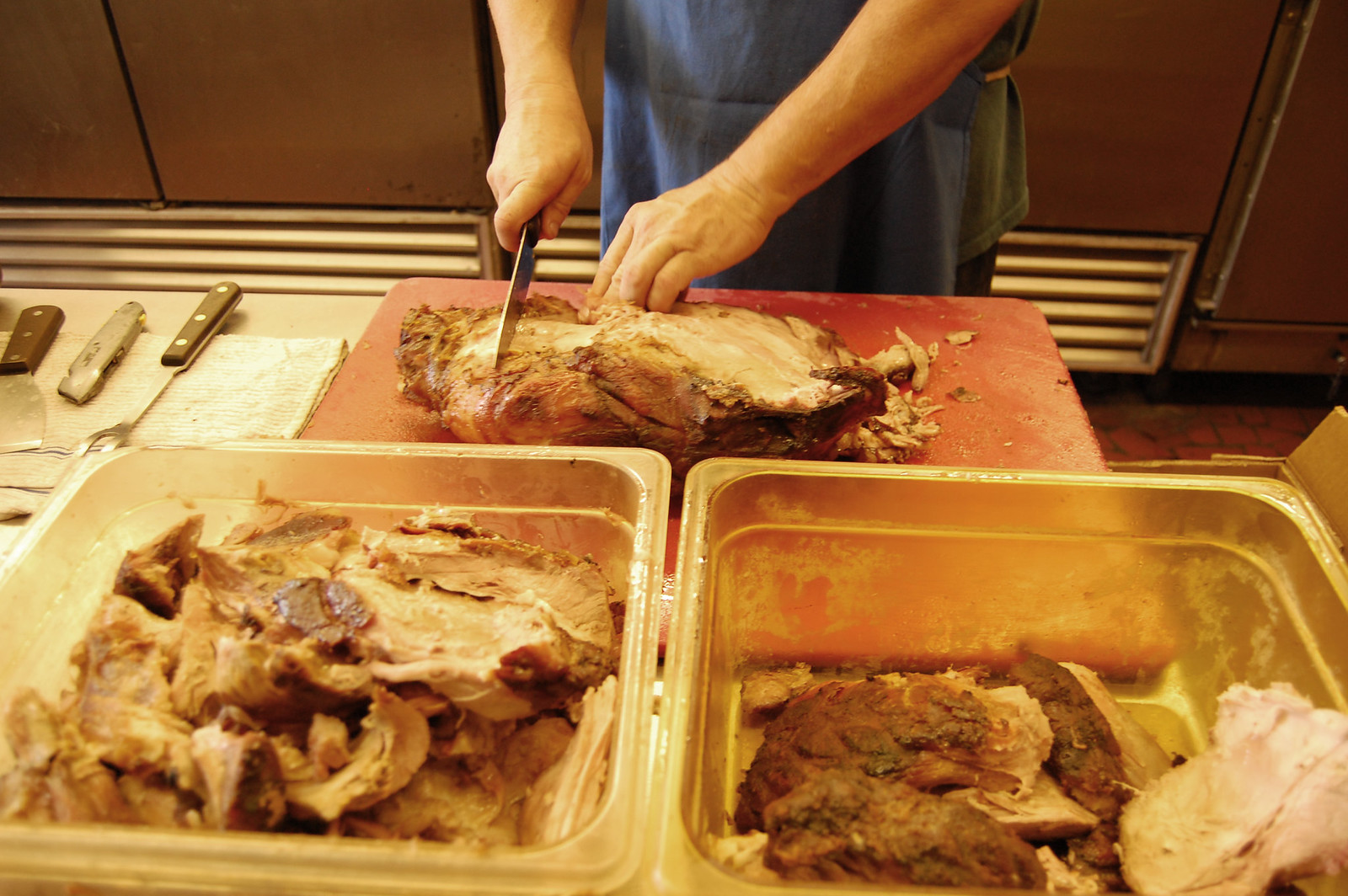 A man in a blue apron works a long knife through a cooked pork shoulder on a red cutting board, holding the meat steady with his other hand; the crusted outside is dark and the cut face is pale. Two steel hotel pans stand in front of him, one heaped with pulled and chopped pork, the other holding larger pieces in their own juices. Two cleavers and a knife lie on a cloth at the left; stainless kitchen equipment behind.
