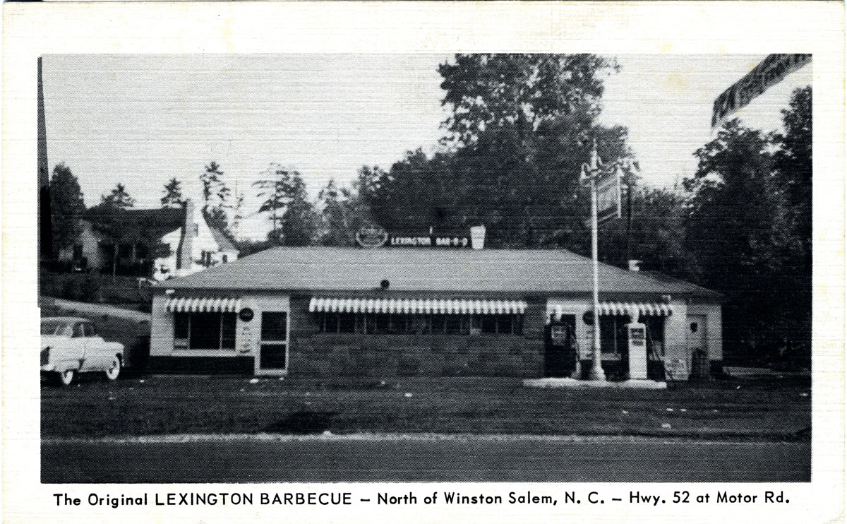 A black-and-white postcard of a single-storey roadside restaurant with a stone-faced lower wall and striped awnings over each window. A sign along the roof ridge reads LEXINGTON BAR-B-Q; a tall pole sign stands at the right edge of the frame, and two petrol pumps and a vending machine sit by the right-hand door. A light-coloured car of the early 1950s is parked at the left. Houses and trees on a rise behind. Printed beneath the image: “The Original LEXINGTON BARBECUE — North of Winston Salem, N. C. — Hwy. 52 at Motor Rd.”