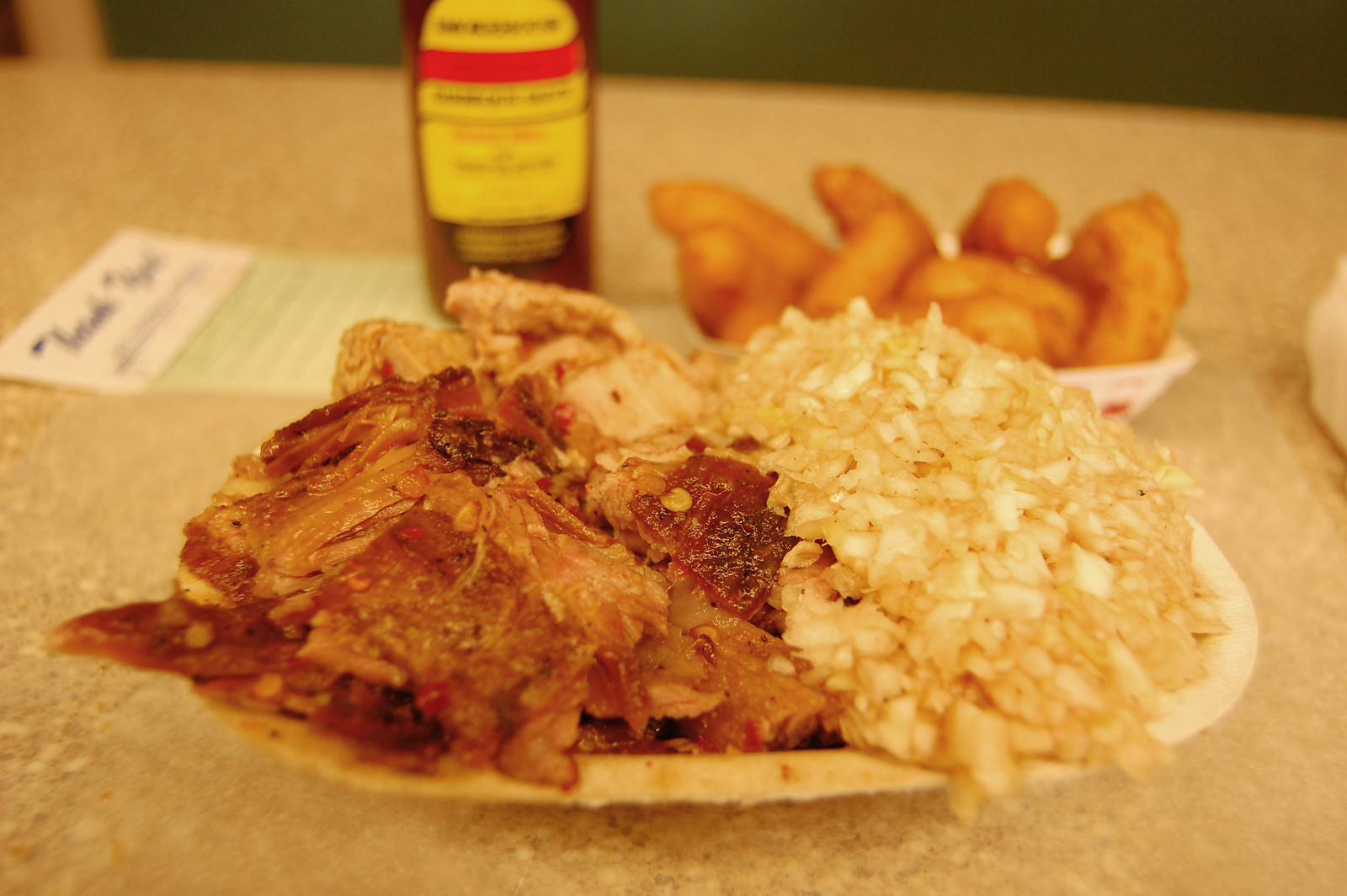 A paper boat on a formica counter holding chopped pork with flecks of dark bark and red pepper through it, and beside it a heaped mound of finely chopped slaw. Behind, a second paper boat of hushpuppies, a bottle of sauce with a yellow and red label, and a printed card.