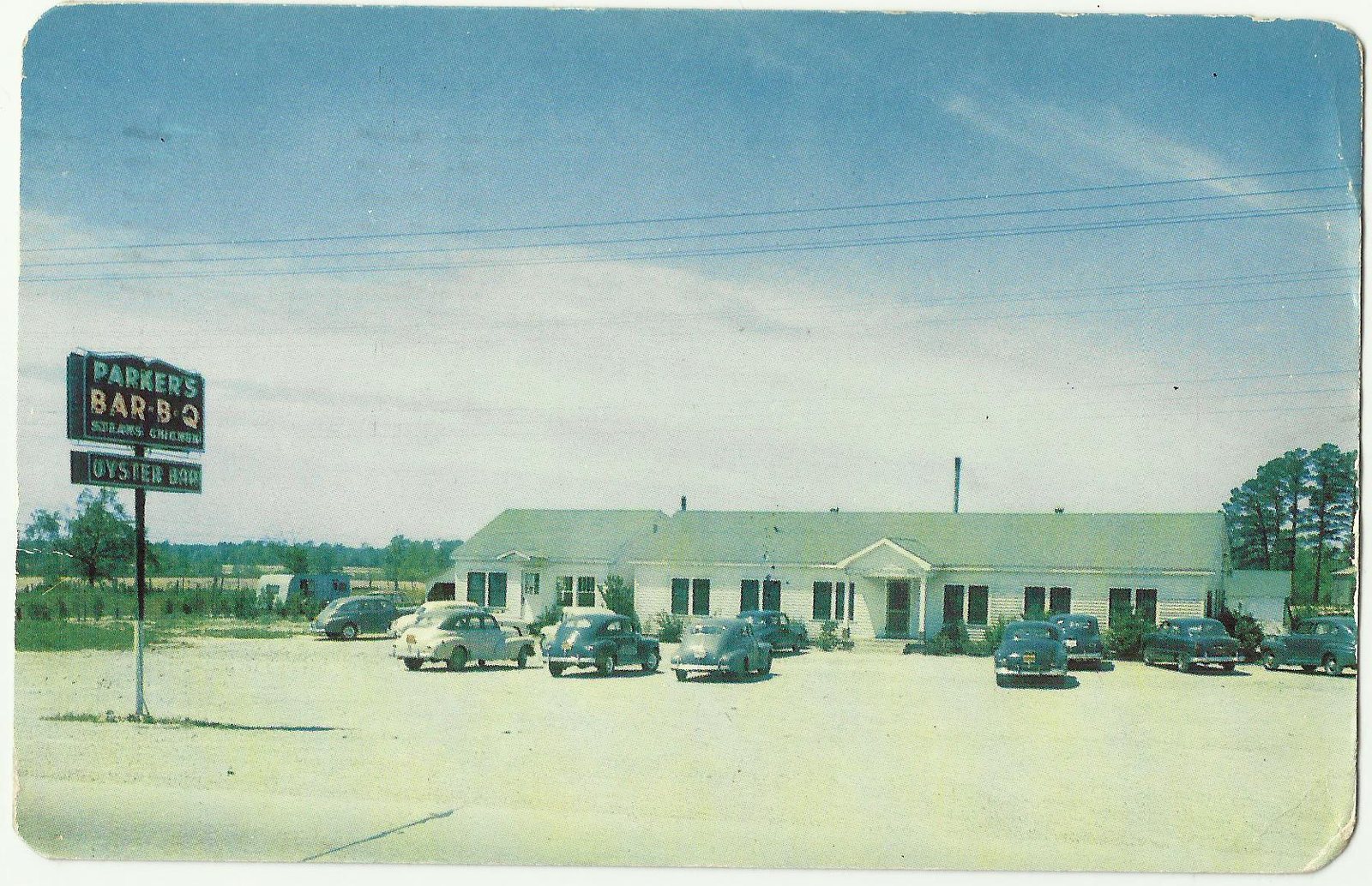 A colour-tinted postcard of a long low white building with a small columned porch at the centre, seen across a bare sand lot with eight or nine cars of the 1940s parked nose-in. A steel post at the left carries a sign in two panels: PARKERS BAR-B-Q over STEAKS CHICKEN, with a smaller board beneath reading OYSTER BAR. Pines and open field run along behind the roofline.