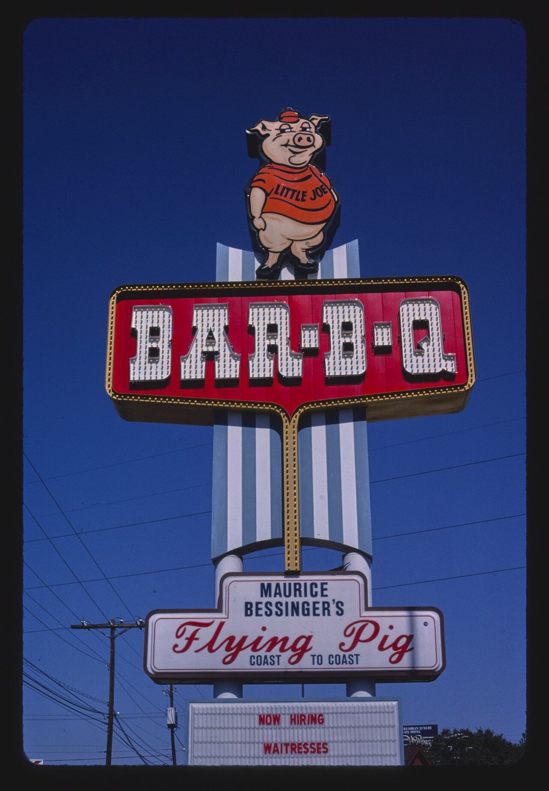 A pink pig in a red shirt lettered LITTLE JOE, cap on its head and hands behind its back, standing on the crown of a tall roadside sign above the words BAR-B-Q. Maurice Bessinger's, West Columbia, South Carolina, 1988.