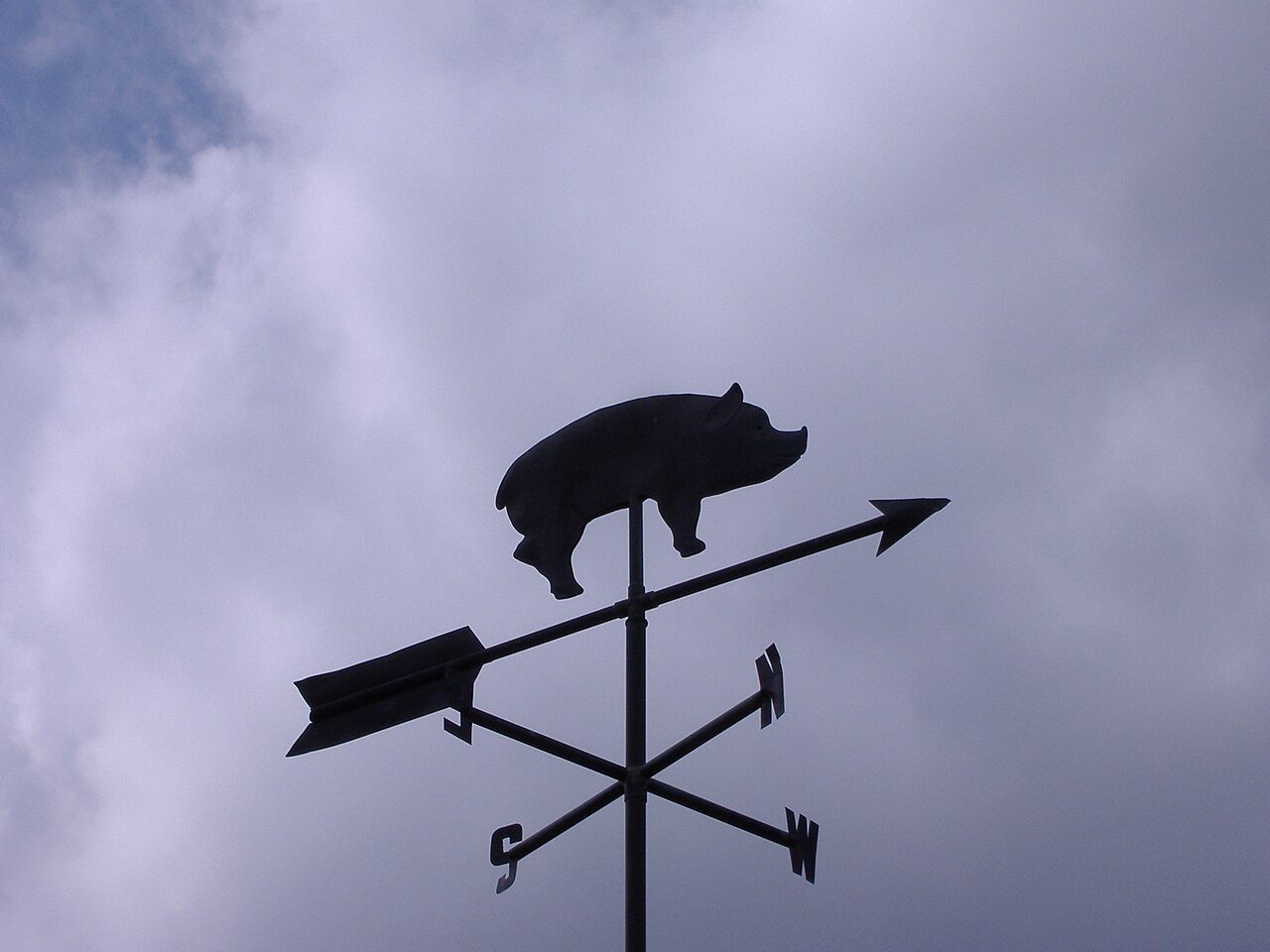 A flat sheet-metal pig cut in silhouette, running nose into the wind on a weather vane above the roof of Wilber's Barbecue, Goldsboro, North Carolina, with the four cardinal letters below it.