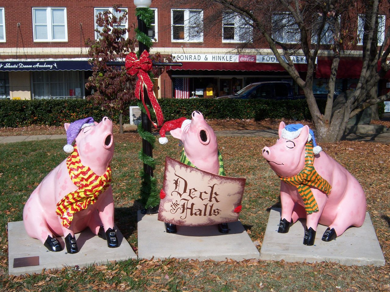 Deck the Halls: three small pink pig sculptures in Santa hats and striped scarves, set around a signboard lettered Deck the Halls on a lawn in front of a street of shops.