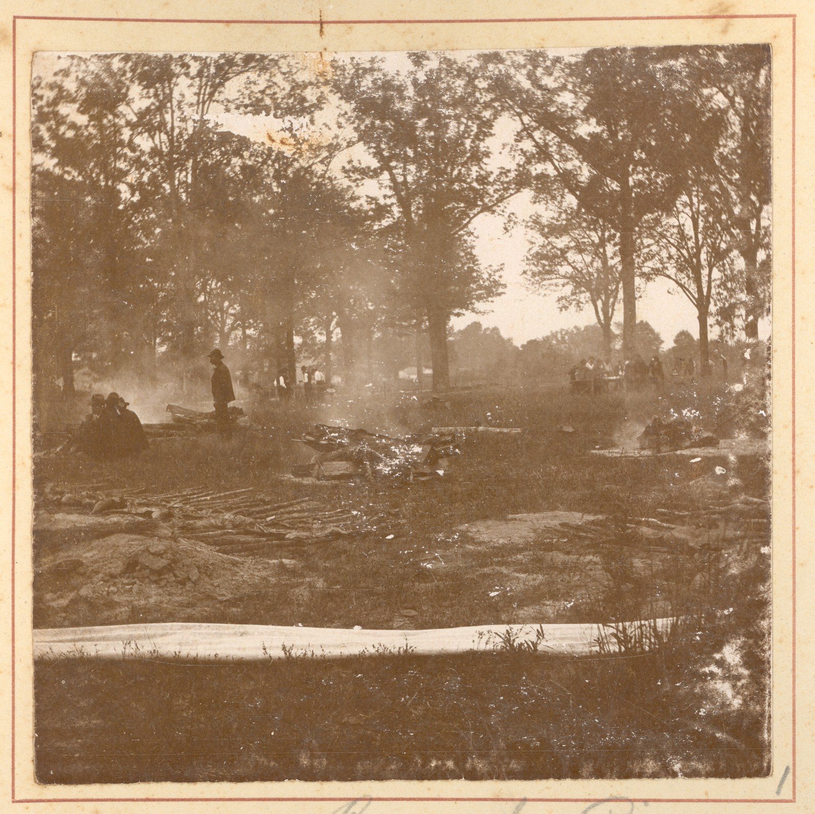 Smoke hangs in the air among tall bare-trunked trees. In the middle of the frame a man in a dark hat and coat stands in profile beside a low line of banked earth with meat and burning wood on it; two people sit on the ground to his left. Further back, small figures move around tables in a clearing. Across the foreground run the low ridges of a dug trench and a band of white canvas laid flat on the grass. Sepia print mounted on card.