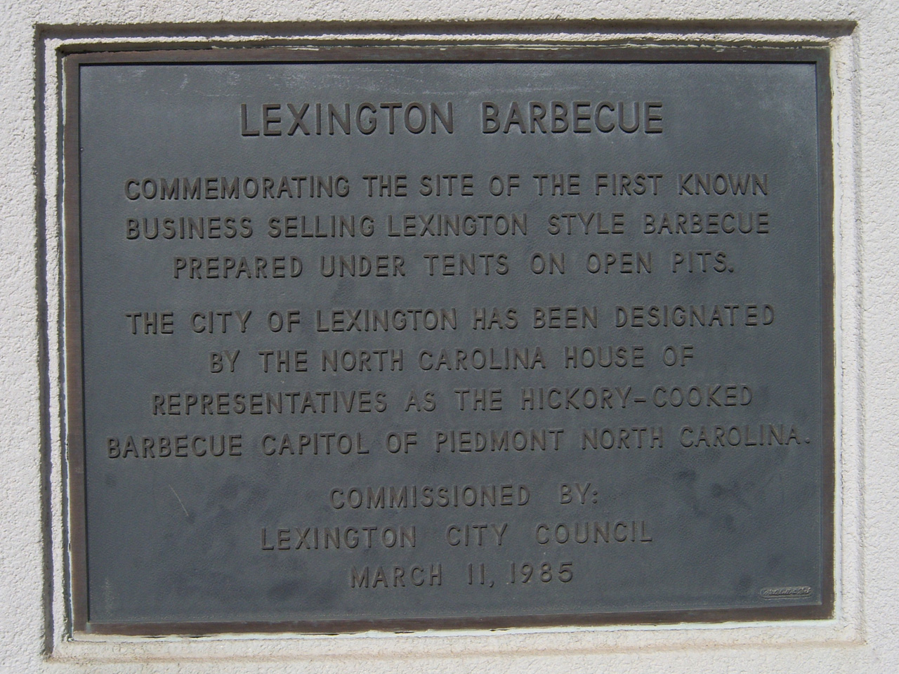 A bronze tablet set into a stucco wall, lettered LEXINGTON BARBECUE, commemorating the site of the first known business selling Lexington-style barbecue prepared under tents on open pits, and recording the North Carolina House of Representatives' designation of the city as the Hickory-Cooked Barbecue Capitol of Piedmont North Carolina. Commissioned by Lexington City Council, March 11, 1985.