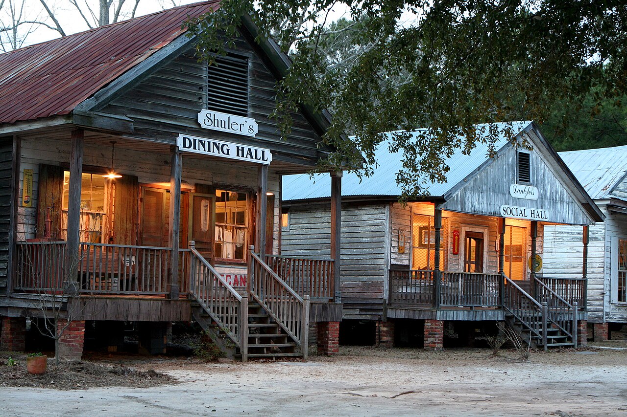 Two old frame buildings on brick piers at dusk, lit from inside, with painted wooden boards hung from the porch roofs: Shuler's DINING HALL on one and Dantzler's SOCIAL HALL on the next. Lone Star BBQ and Mercantile, Santee, South Carolina.