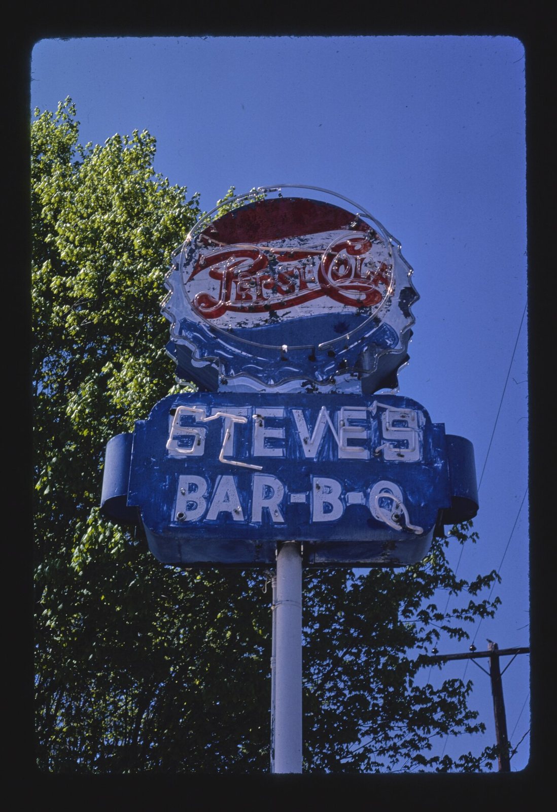 A blue steel sign cabinet on a pole with STEVE'S and BAR-B-Q in white neon on two lines, and above it, larger than either, a chipped Pepsi-Cola bottle cap in red, white and blue neon. Spencer, North Carolina, 1982.