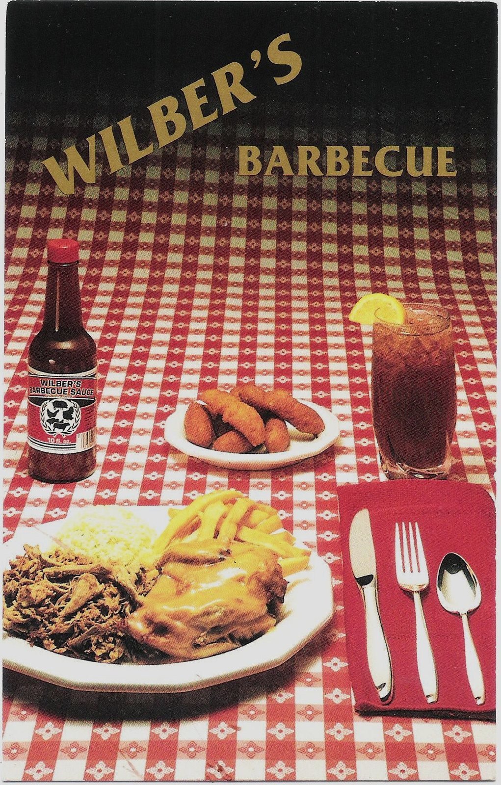 A colour postcard of a meal laid on a red-and-white checked tablecloth, the words WILBER'S BARBECUE across the top in gold. On the large plate, a mound of chopped pork, a scoop of pale slaw, french fries and a piece of chicken under gravy. Beside it a side plate of hushpuppies, a tall glass of iced tea with a lemon wedge, a bottle of Wilber's barbecue sauce with a pig on the label, and a knife, fork and spoon laid on a red napkin.