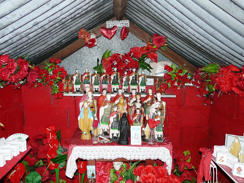Inside a red Réunion chapel: rows of Saint Expedite statues among red flowers.