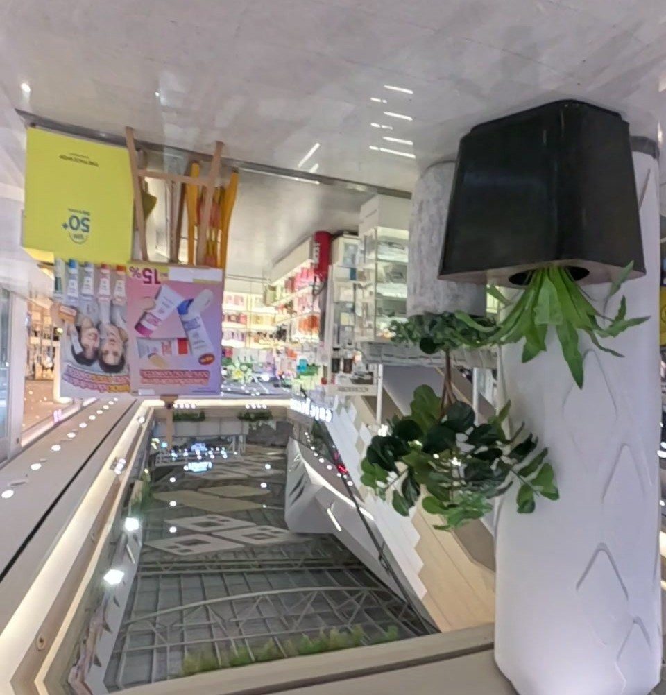 Atrium void seen from an upper floor: escalators drop to the skylit ground floor, a cosmetics shop and hanging plants alongside.