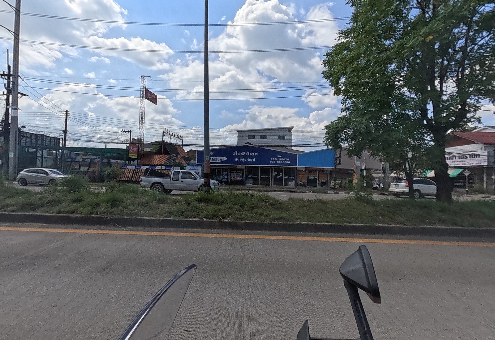 Across the median, a blue Samsung shop signed วีไอพี บีเทค ศูนย์สุขภัณฑ์, a roof-tile yard with a 089-4810401 pole sign, and a white pickup.