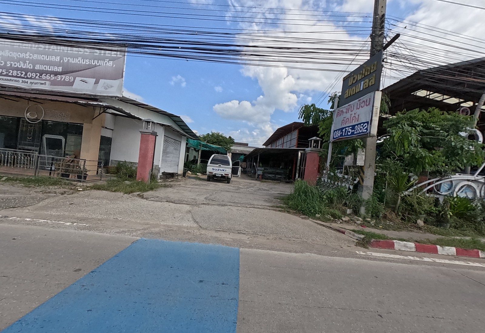 The yard of ม่วนไทย สแตนเลส with its pole sign ตัด พับ ม้วน ดัดโค้ง, a stainless-steel billboard over a closed shop, a white Suzuki van in the drive and curved steel pieces on display.