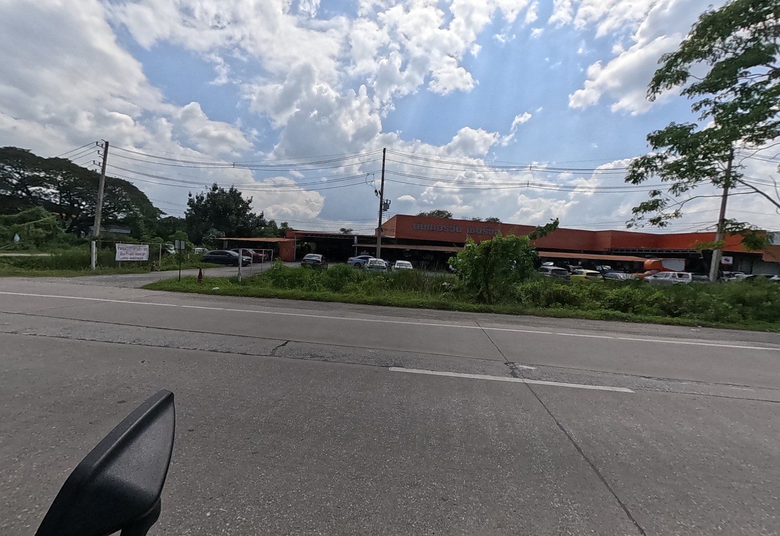A long orange garage building with cars parked in front sits behind a scrubby verge; a white banner hangs on a fence by its gravel drive.