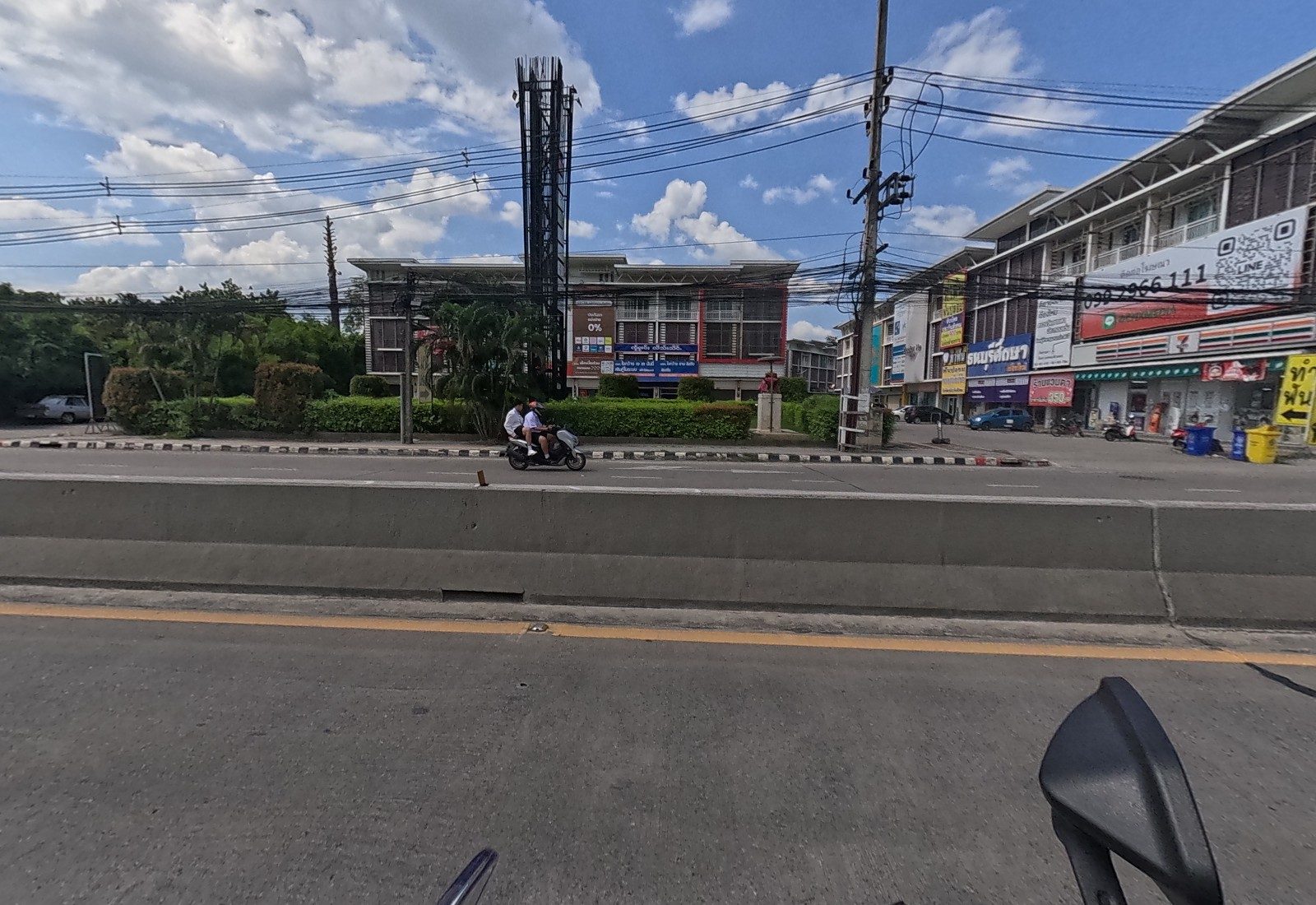Across the road: a shophouse row with a 7-Eleven, 'ธนบุรีศึกษา', a massage shop and yellow 'ทำฟัน' dental arrows, next to a steel sign tower and a building with a 0% loan ad.