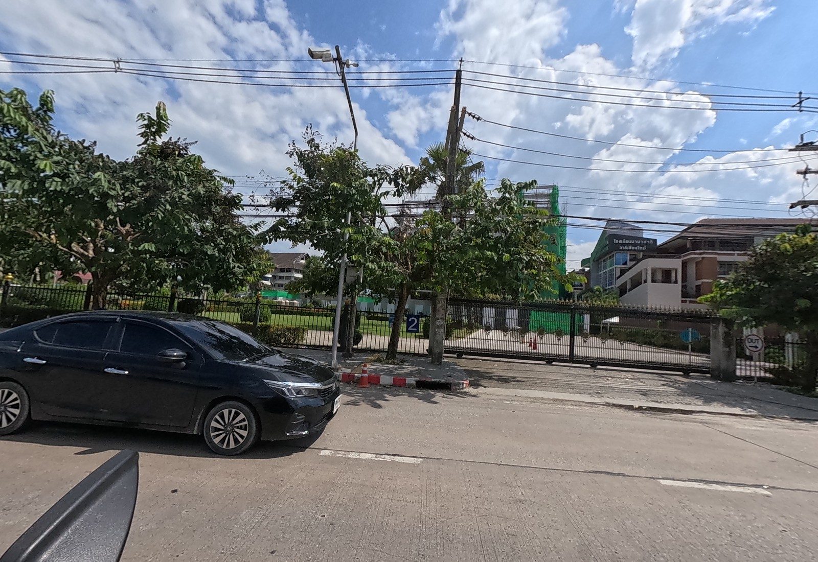 A black saloon passes a school gate numbered 2 with an OUT sign; behind the fence, the netted building under construction and a green-signed school block.