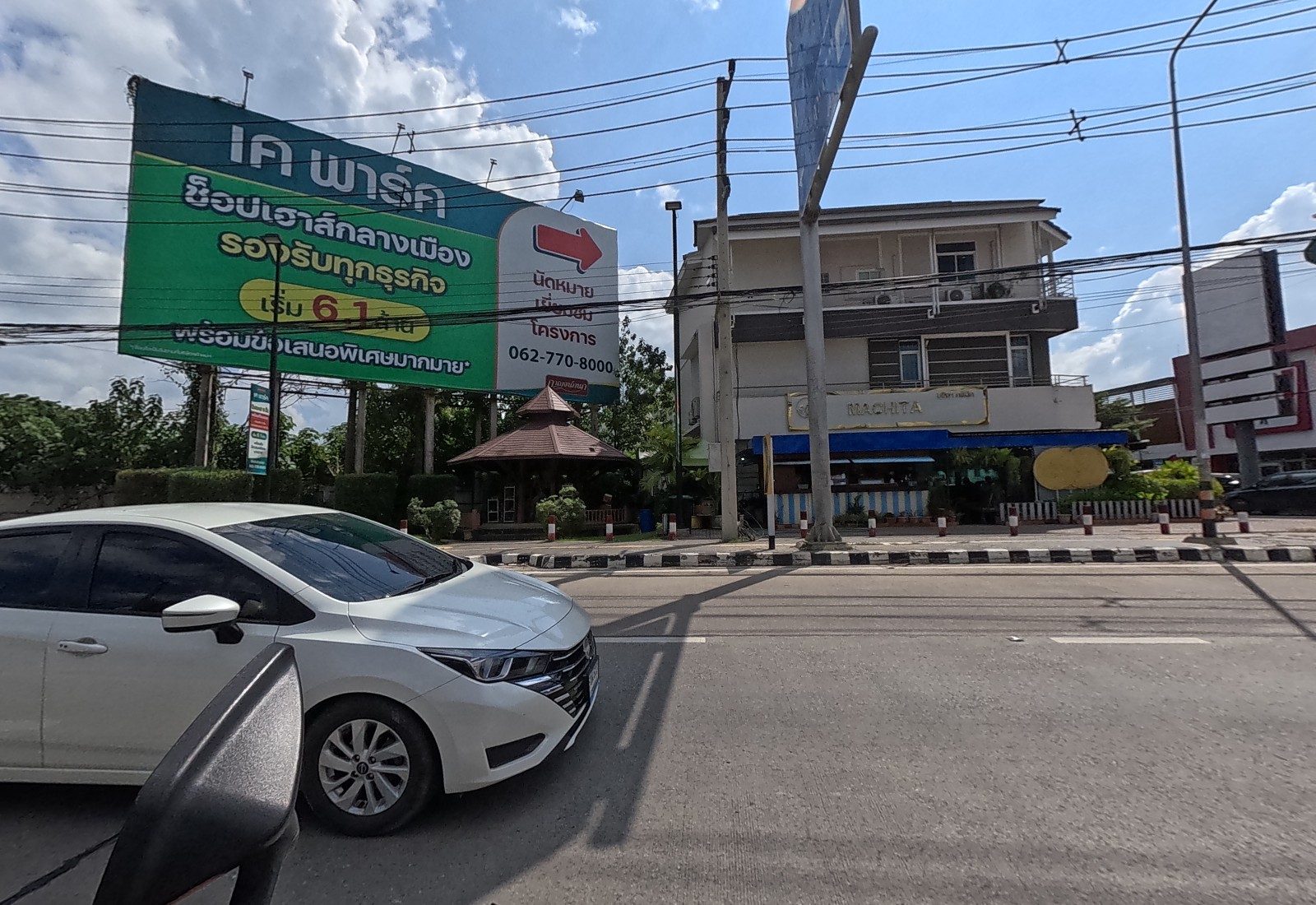 A big green billboard for the K Park shophouse project with a teak pavilion, and a three-storey building with the Machita sign; a white car alongside.