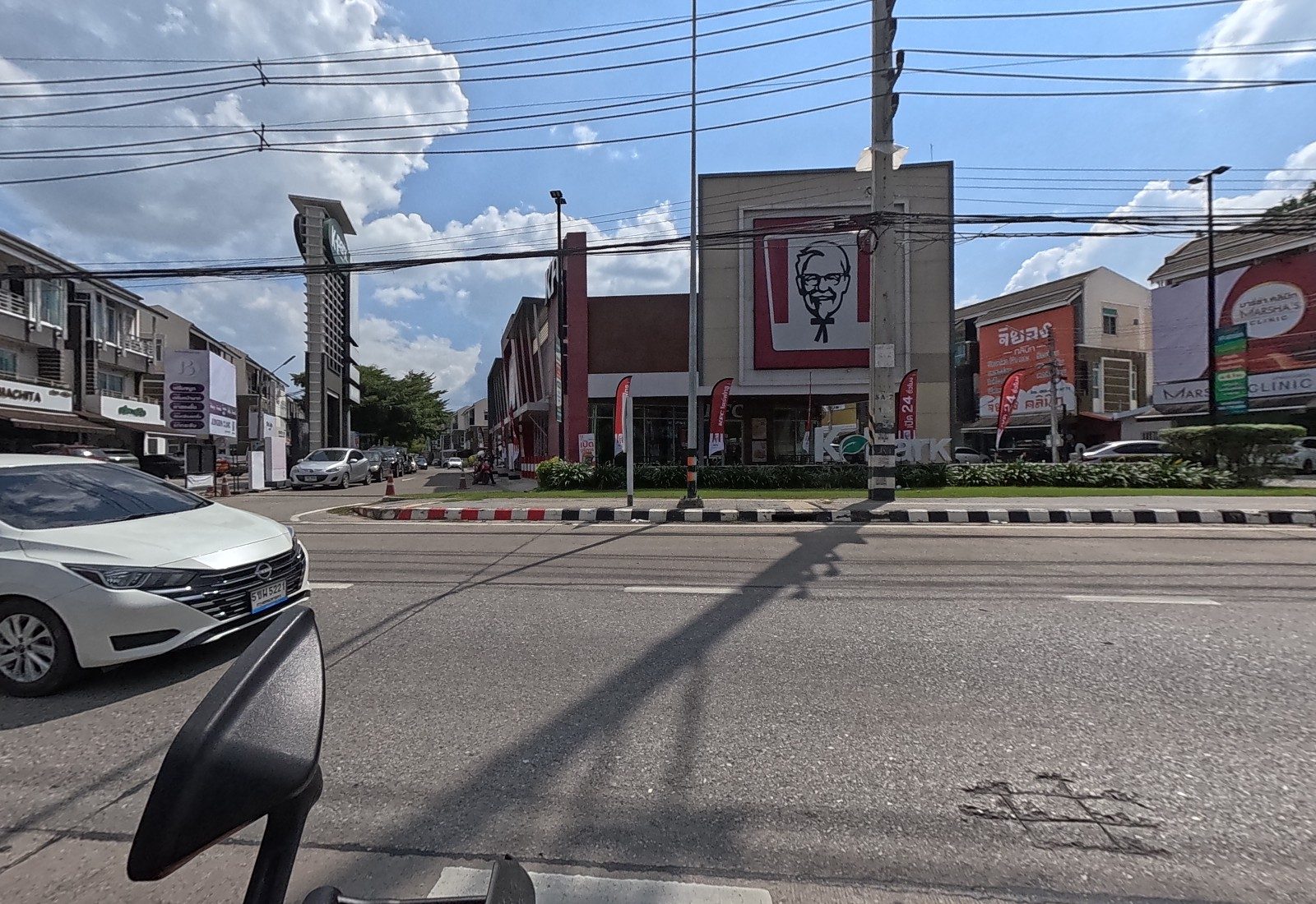 The K Park shophouse estate entrance: Machita, a small shop and a Longen Clinic sign on the left, the KFC building in the middle, clinic signs on the right.