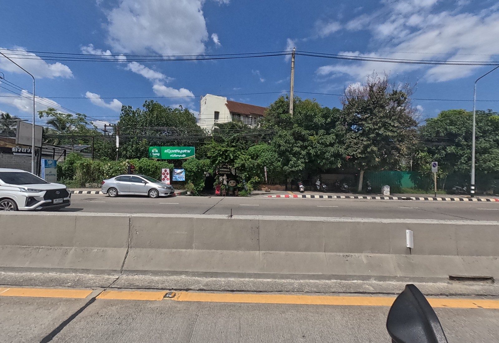 Across the road, a garden restaurant under trees with a green Chang sign reading ครัวเพชรดอยงาม, a mascot figure at the gate, and a car-wash building to the left.