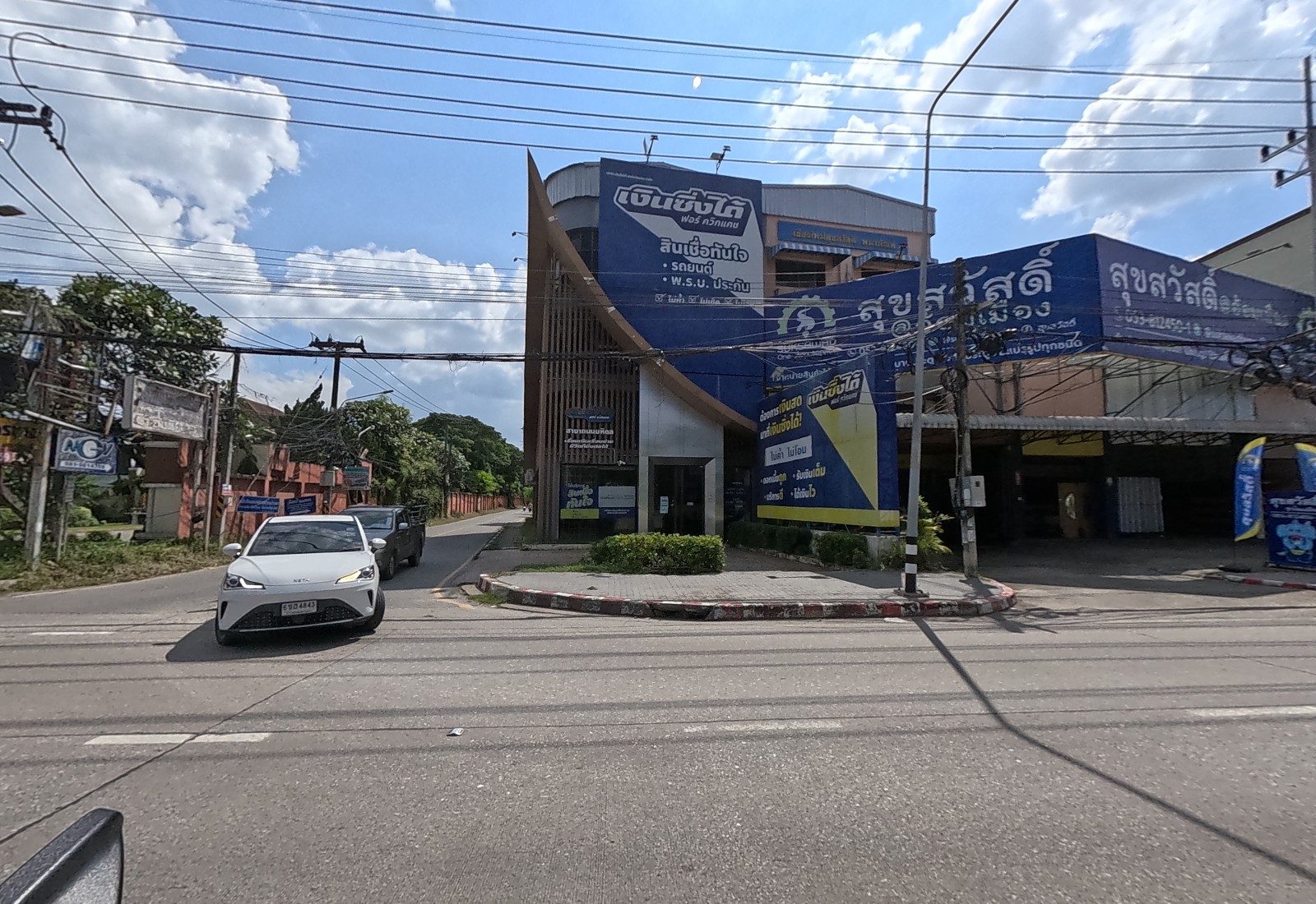 A soi opens beside a blue-signed Ngern Sing Dai loan office and the Suksawad timber shop, with a white car coming out.