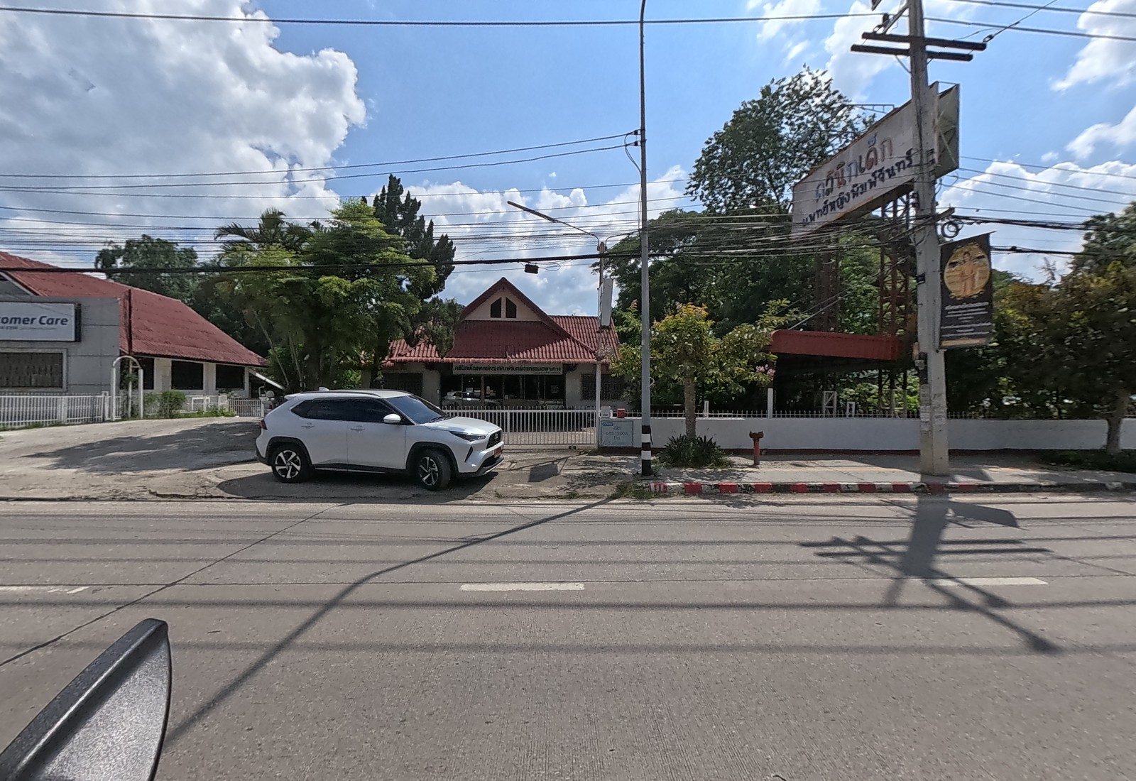 A white SUV is parked in front of the children's clinic, a red-roofed house with a tall pole sign, beside the Beko office.