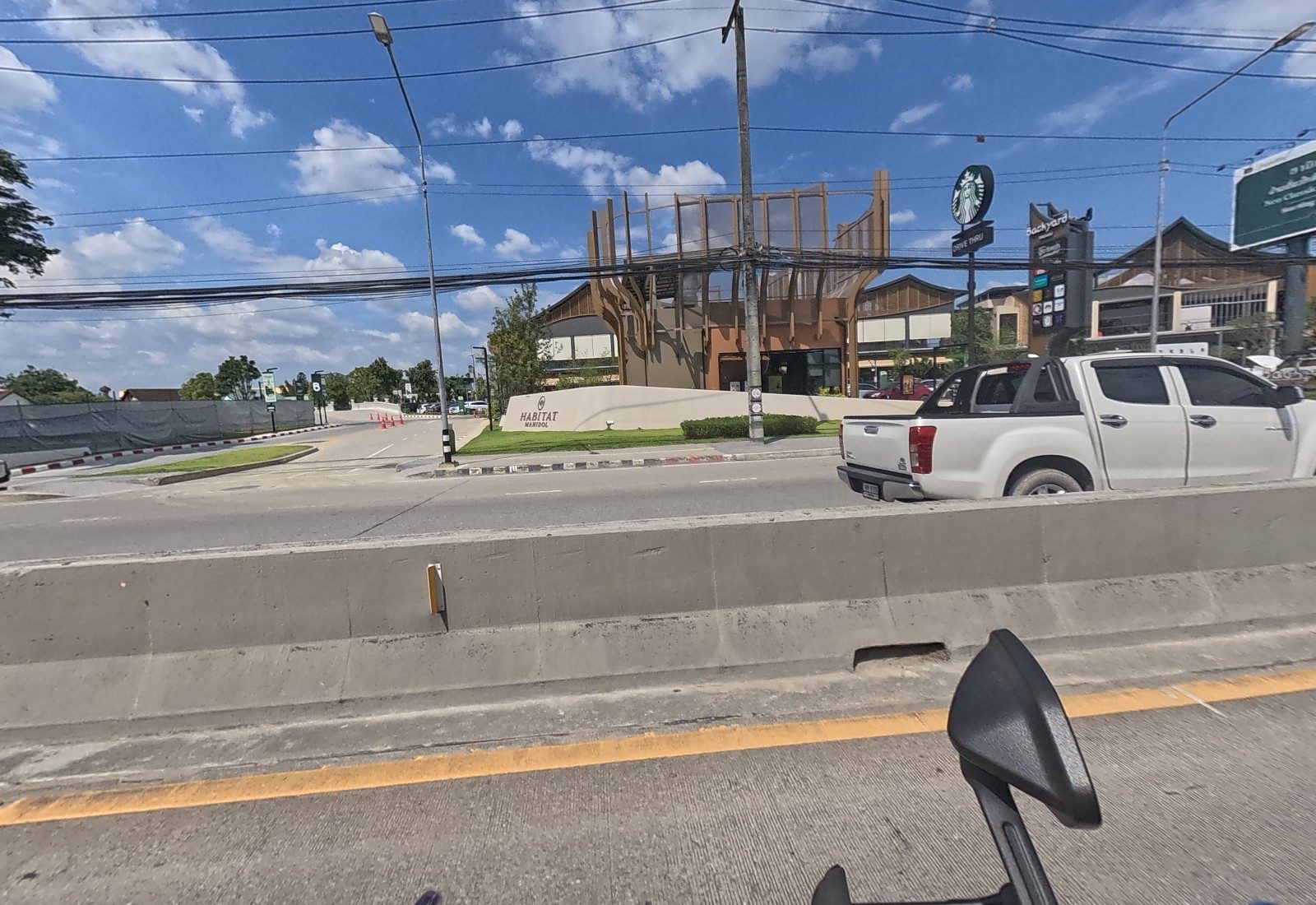 Across the median, the Backyard mall pylon, the Starbucks drive-thru sign and the Habitat Mahidol entrance wall stand behind passing pickups.