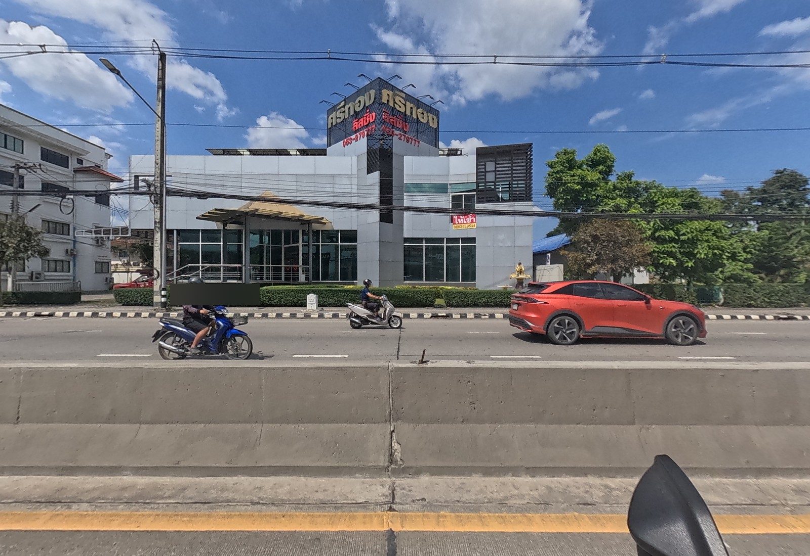 Across the road, a grey showroom with a gold canopy and a rooftop sign for ศรีทอง ลิสซิ่ง, posted for rent, with a spirit house beside it.