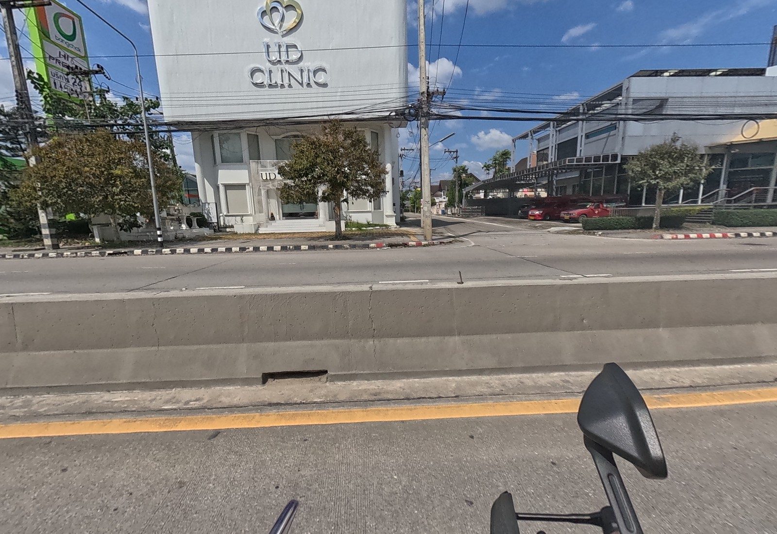 Across the road, the white UD Clinic building at a side-road corner, with a Bangchak pole sign at left and two red songthaews parked by a grey building.