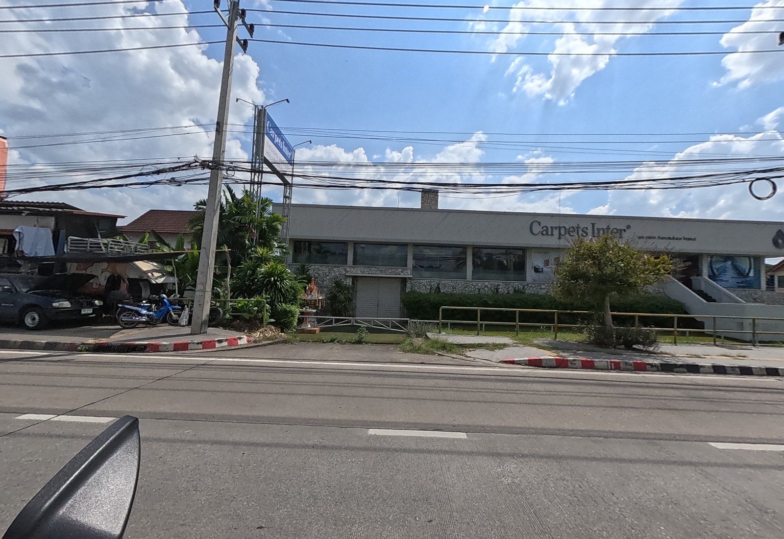 The low grey Carpets Inter showroom with a spirit house at the gate, beside a yard of parked cars and motorbikes.