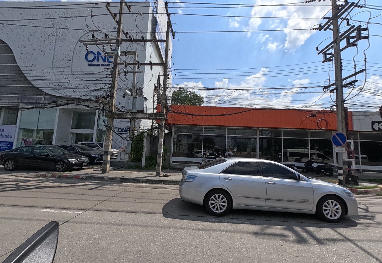 The ONE Medical Clinic building and a long orange-roofed glass-fronted gym with a no-parking sign; a silver car passes.