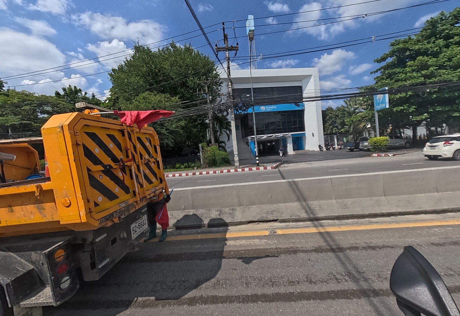 A road-works truck with a flagman beside the median; across the road, the white Krungthai Bank branch with ATMs and parked motorbikes.