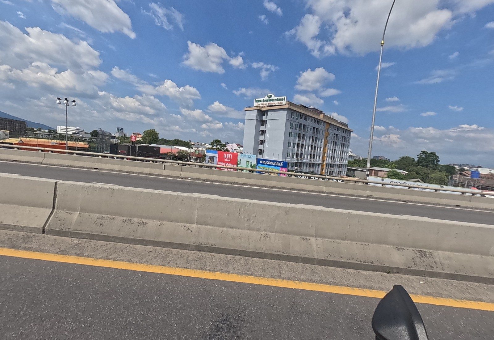 Across the median, The Airport Greenery Hotel rises behind a row of housing-project boards including Kanok Ville.