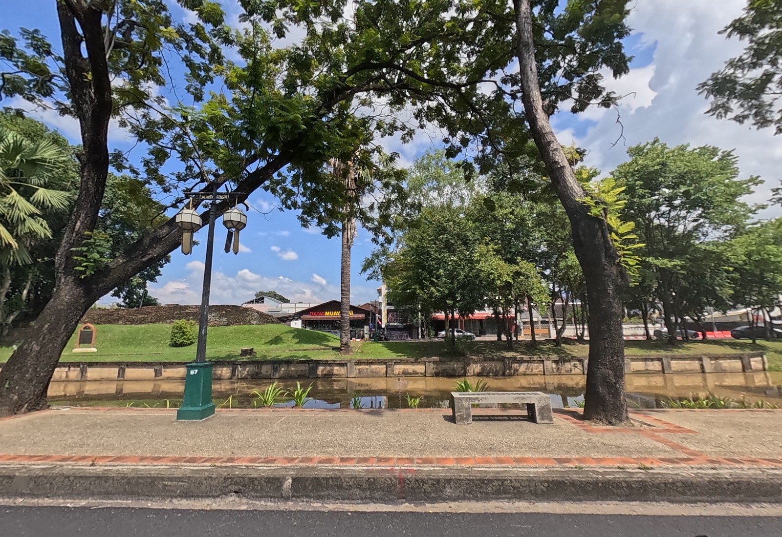 Across the moat, a grassy stretch of the old city wall with a plaque, and the Dang Muay Thai building beyond; a stone bench and lamppost 87 on the footpath.