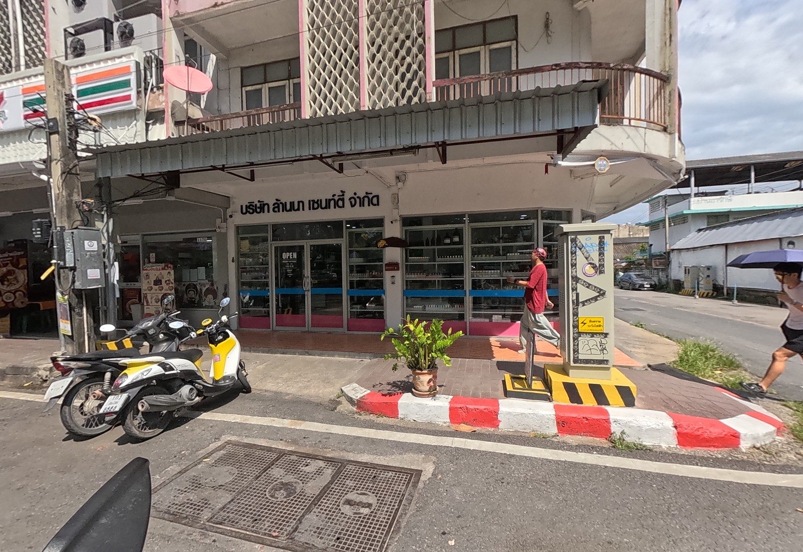 The full corner front of บริษัท ล้านนา เซนท์ตี้ จำกัด with a man in red walking past and a pedestrian with an umbrella crossing the side street.