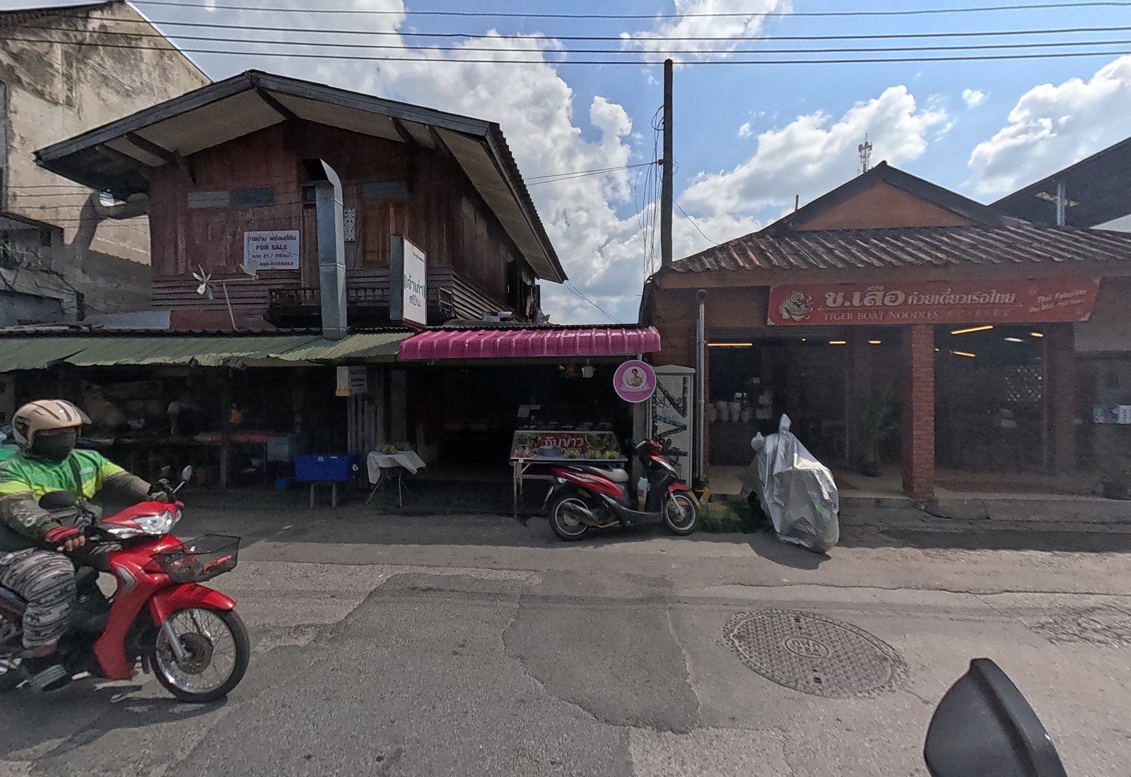 An old wooden house with a FOR SALE sign over a pink-awning rice-and-curry stall, next to the Tiger Boat Noodles house; a Grab rider passes.