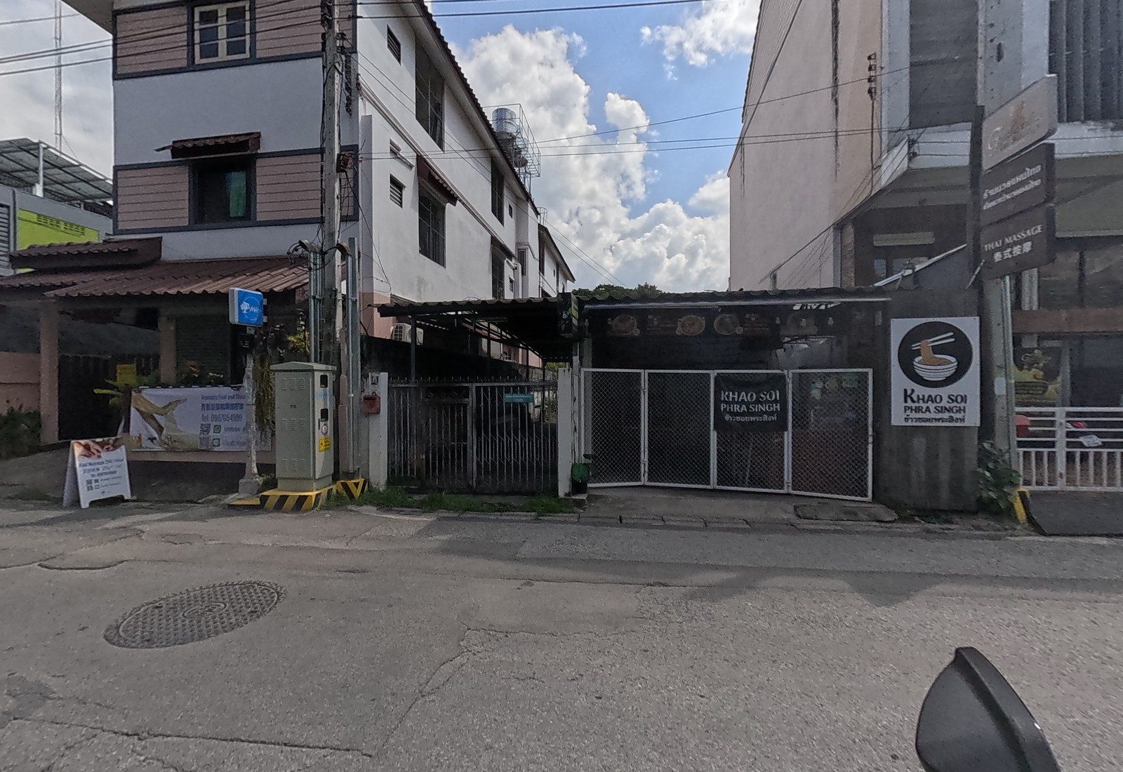 Khao Soi Phra Singh sits behind a closed mesh gate with a bowl logo sign, beside Getthawha's hanging signs and a lane with a foot massage banner and board.