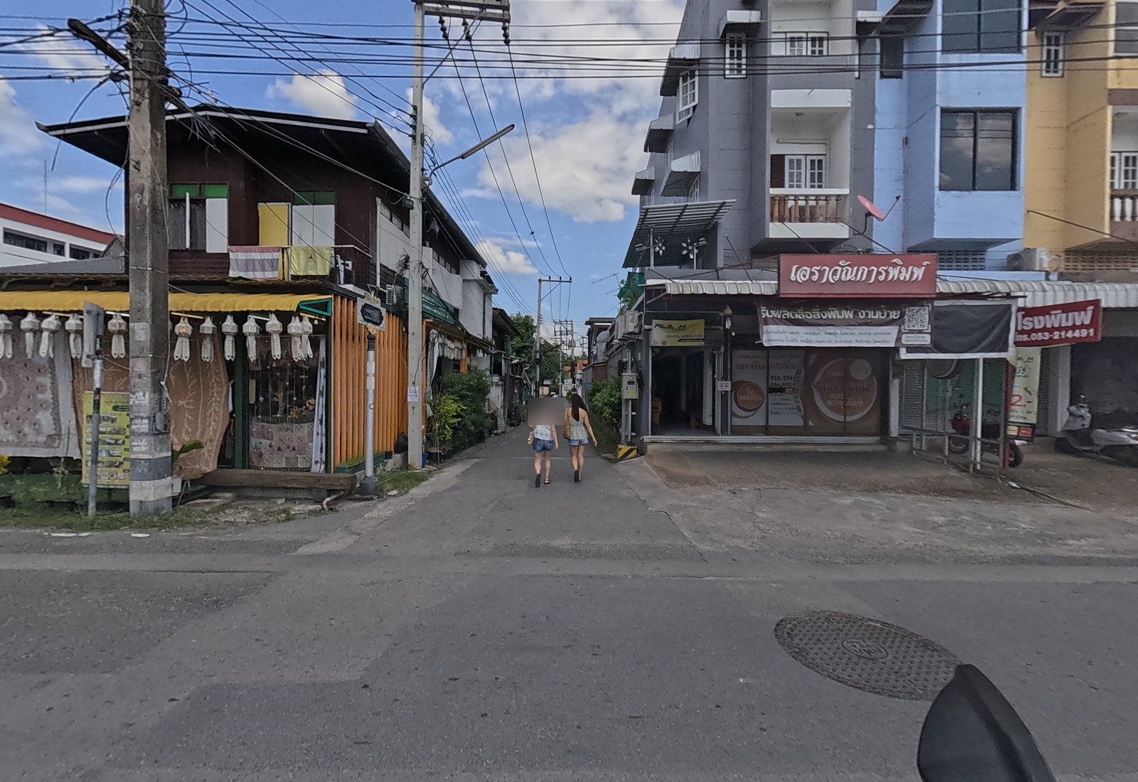 Two women walk down a lane between an old teak house hung with white lanterns and textiles and the เอราวัณการพิมพ์ printing shop with a FLASH sign.