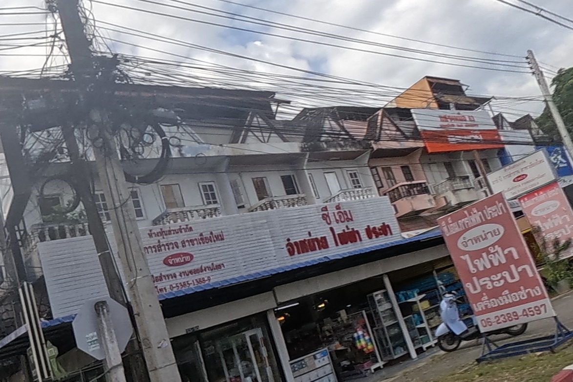 A hardware shop selling electrical and plumbing supplies sits on the corner with a red freestanding sign and phone number, and the road continues past parked cars toward a pedestrian and more shops.