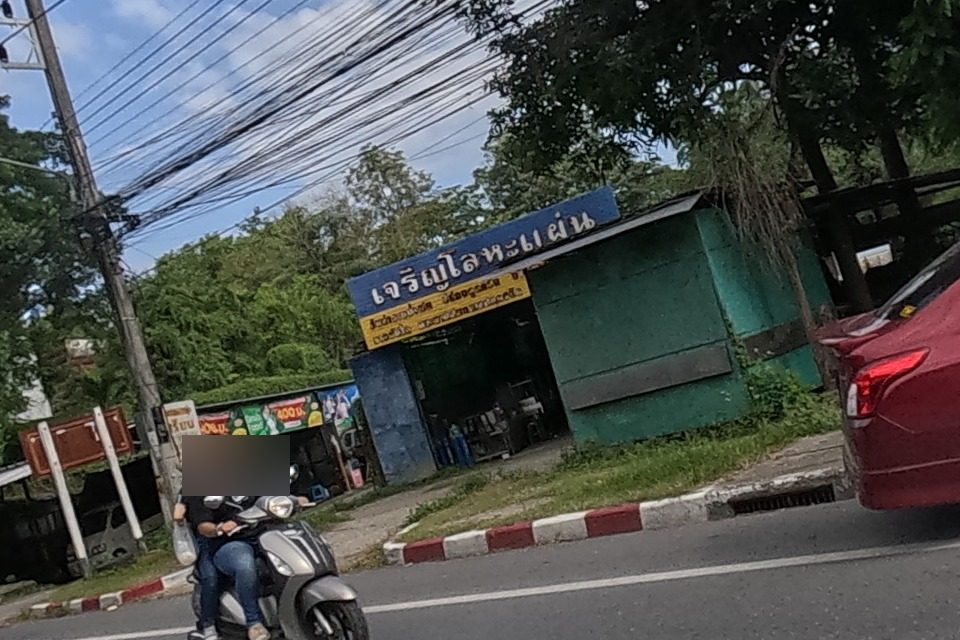 Across the road, a sheet-metal fabrication shop with a green and yellow sign sits beside a car-battery banner ad; a woman rides a scooter past and a red sedan is turning.